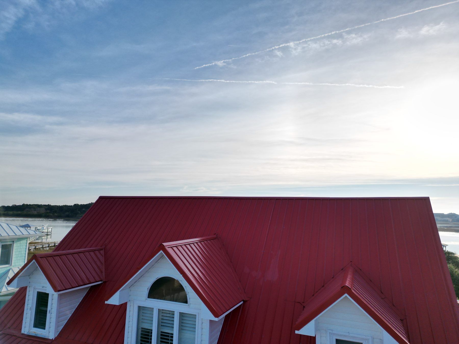 Red standing-seam metal roof on a white coastal house, with water and sky behind