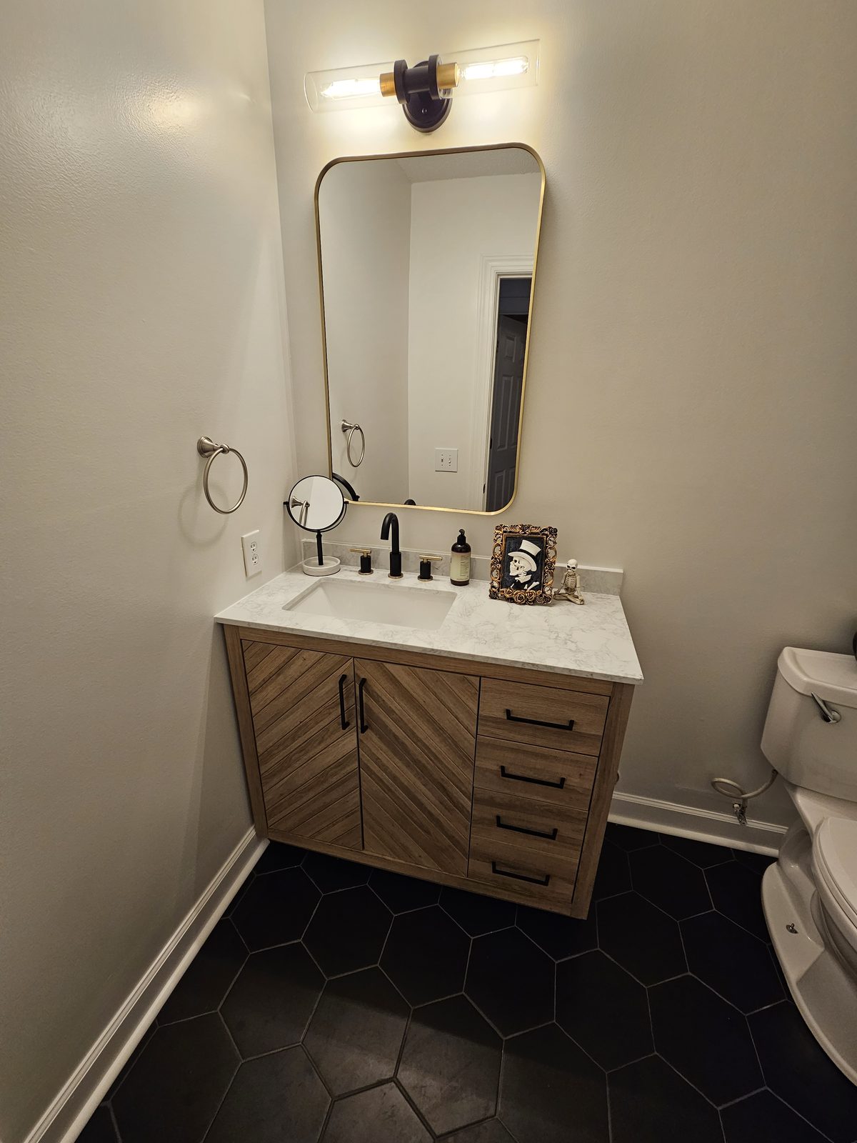 A wood vanity with a round mirror over a black hexagon tile floor