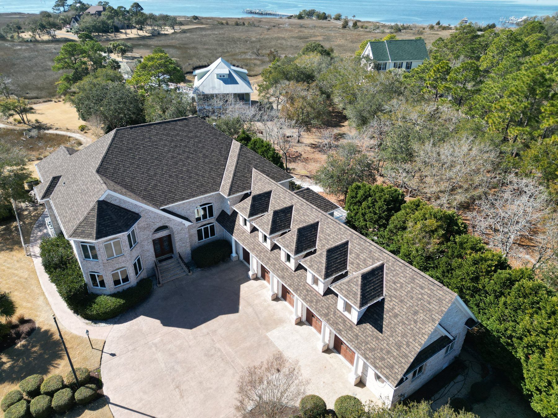 Aerial view of a large brick home with a new-looking shingle roof and a row of dormers, beside coastal marsh and open water